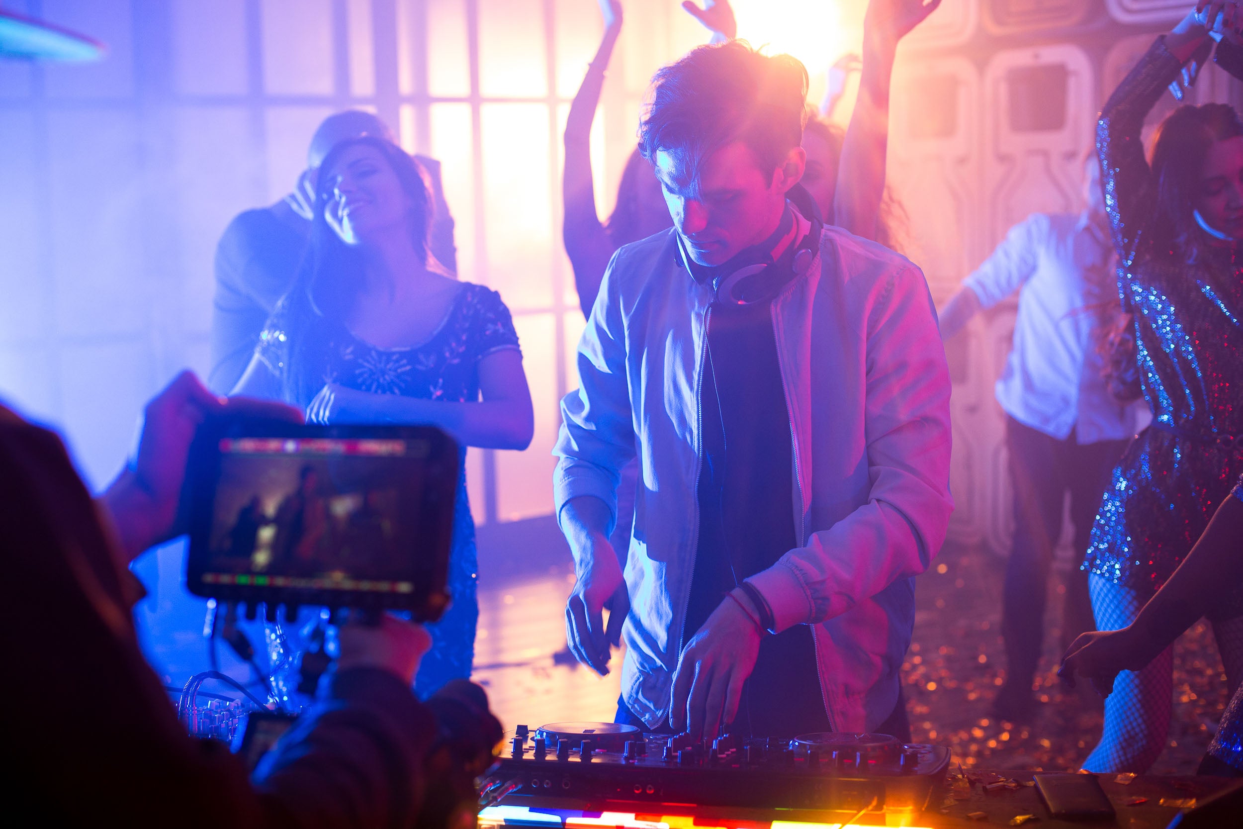 DJ at a party, with people dancing around him and somebody is filming him, while he uses the DJ mixer deck. 